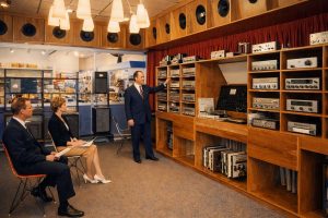 1970s hi fi showroom with salesman demonstrating stereo receivers and speakers to seated customers